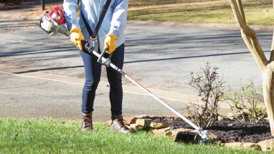 Wide view of person using a handheld trimmer to trim the edges of a lawn around a flowerbed.  