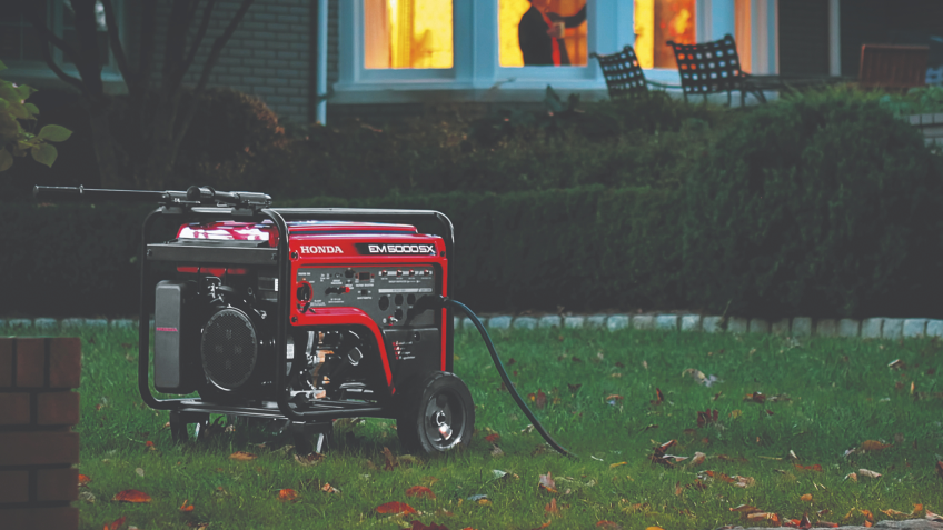 Une génératrice Honda EM5000 rouge sur une pelouse à l'extérieur d'une maison en brique bordée d'arbustes.