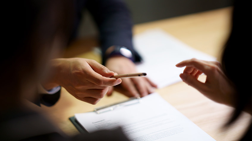 Close-up of a person passing a pen to another person who has documents on the table in front of them. The edges of the photo are blurry, with the emphasis only on swapping the pen between the hands.