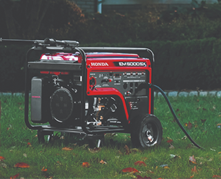 Une génératrice Honda EM5000 rouge sur une pelouse à l'extérieur d'une maison en brique bordée d'arbustes.